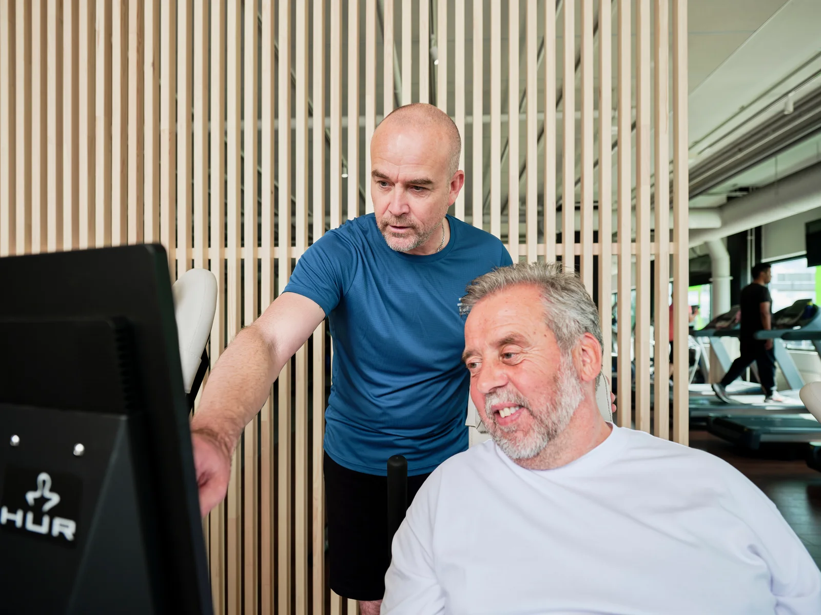 An instructor beside a seated exerciser at a HUR touchscreen
